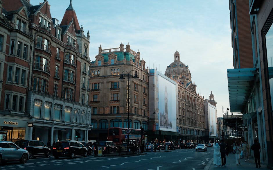 A city street scene featuring historic Victorian-style buildings with ornate architectural details, including a prominent domed structure in the background. The street is bustling with cars parked alo